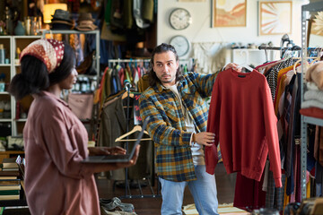 Caucasian young adult man showing sweatshirt to Black young adult woman while shopping in thrift shop, man holding clothing on hanger and woman gesturing with hands, racks of clothes in background