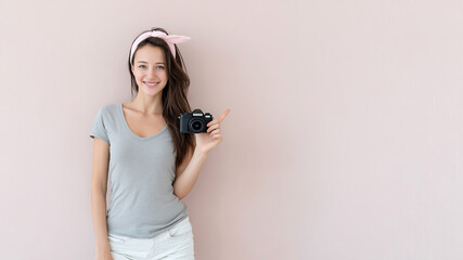 A young beautiful Asian girl with a camera in her hand on a pink background points to a free space where your text can be placed.