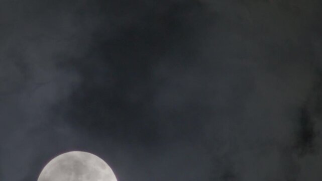 The full moon slowly ascends through dark clouds in a moody night sky, marking the quiet onset of nighttime.