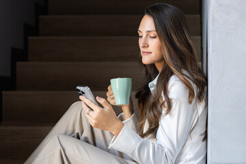 A serene young woman sits on modern stairs, engrossed in her smartphone while enjoying a warm beverage