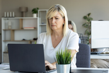 A diligent blonde businesswoman works intently on her laptop in a bright, modern office setting