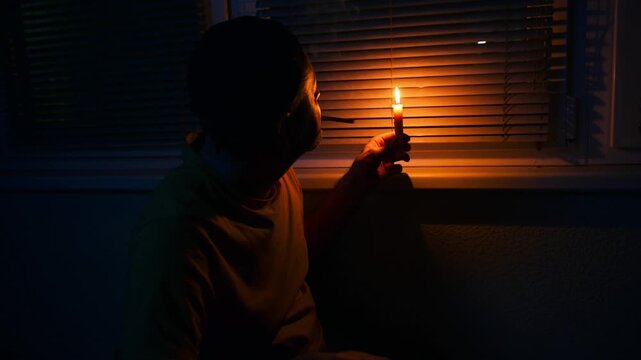 Blackout, No electricity. Man in a dark room near a window with candle. Depressed young man during a power outage.