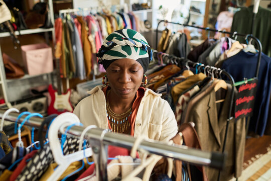 Young adult Black woman browsing clothing rack in thrift shop, selecting garments among assorted secondhand clothes, focused expression, surrounded by vintage apparel displays - Powered by Adobe