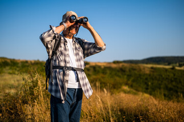 Fototapeta premium Senior man with backpack using binoculars while hiking in nature. 