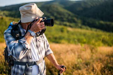 Active senior hiker using camera for photographing while hiking with backpack and hiking poles in nature.	