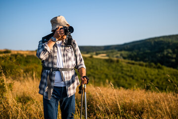 Active senior hiker using camera for photographing while hiking with backpack and hiking poles in nature.	