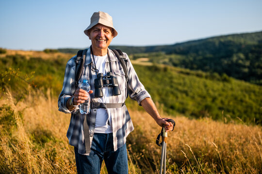 Portrait of happy senior man drinking water while enjoying hiking with backpack and hiking poles in nature.