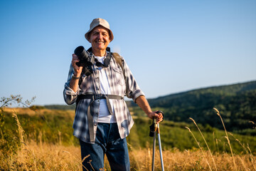 Portrait of happy senior hiker using camera for photographing while hiking with backpack and hiking poles in nature.	