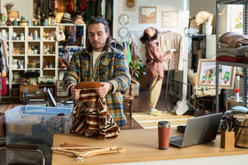 Caucasian young adult man folding patterned sweater at counter in thrift shop, multiethnic young adult woman refreshing clothes with steamer in background, laptop and hangers on desk