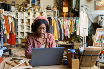 Young adult Black woman working on laptop at desk in thrift shop, surrounded by clothing racks and accessories, focusing on screen with smartphone and hangers nearby