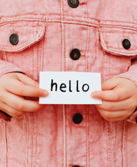 Child's hands holding a white card with the handwritten word "hello" in a pink denim jacket.
