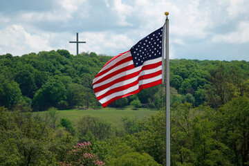 American flag waving on a pole with a large Christian cross on a forested hill.