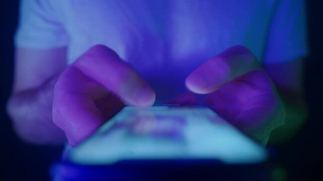 Close-up of hands holding a smartphone and quickly scrolling through social media feeds at night in neon light. Doom scrolling, phone overuse and digital addiction concept. 