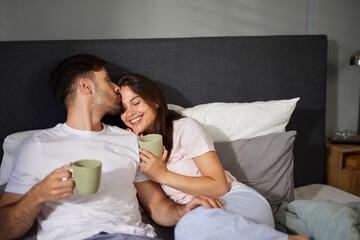 A couple is nestled in bed, sipping coffee from green mugs, sharing joyful moments and smiles in a warm, intimate setting during a weekend morning.