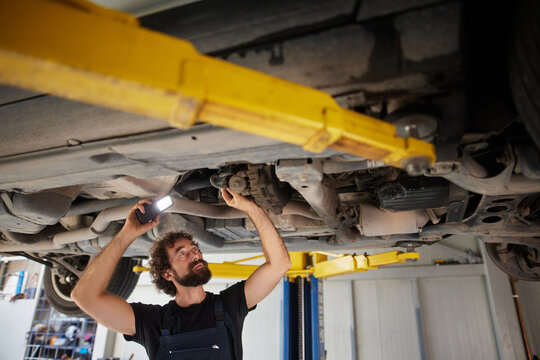 A mechanic works on a car lifted in an auto shop, using a flashlight to examine components beneath the vehicle while focused on the task at hand.