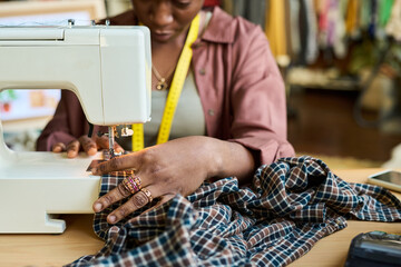 Black woman sewing plaid shirt with sewing machine in thrift shop, hands guiding fabric while measuring tape hangs around neck, focusing on garment repair and upcycling