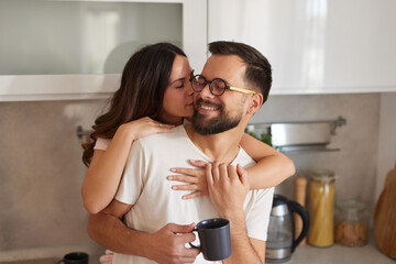 A couple stands in their cozy kitchen, sharing an intimate moment together. They smile and embrace,...