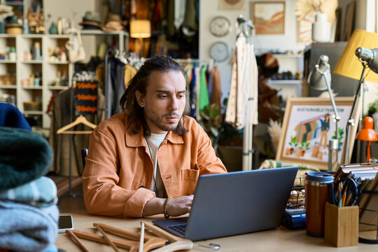 Caucasian young adult man sitting at desk using laptop, surrounded by clothing racks and secondhand items in thrift shop, focusing on screen while working or managing inventory