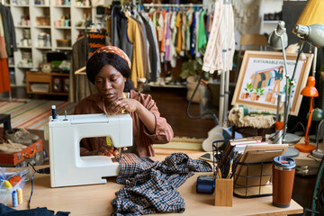 Black young adult woman using sewing machine repairing clothing at wooden table in thrift shop, surrounded by racks of secondhand garments and various office supplies