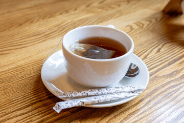 Cup of black tea with tea bag on saucer with spoon and sugar sticks on wooden table.