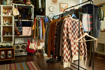 Clothing rack displaying assorted vintage garments in thrift shop, various sweaters and jackets hanging on hangers, retro guitar and stereo equipment visible in background