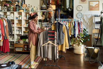Black young adult woman browsing clothing rack in thrift shop, selecting vintage garments surrounded by assorted apparel and accessories, standing near shelves filled with items