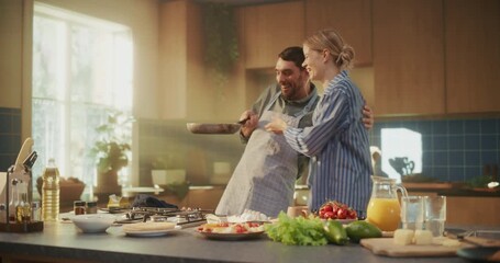 Domestic Lifestyle Scene of a Man Cooking Pancakes in the Kitchen Together with His Girlfriend. He Tosses a Pancake on the Pan while His Partner in Pajamas Stands Nearby, Smiling and Talking to Him - Powered by Adobe