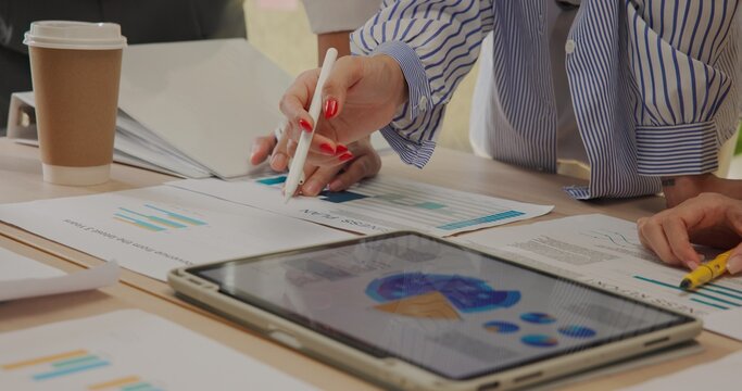 Close-up view of diverse young business team collaborating with digital tablet and printed charts. Hands pointing at colorful graphs and analytics during a strategy planning session at office.