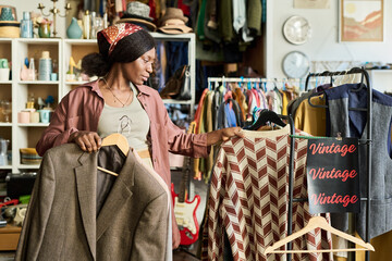 Young adult Black woman examining vintage clothing rack in thrift shop, holding patterned sweater and blazer while looking to side, surrounded by assorted retro garments and accessories