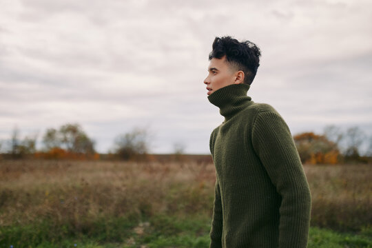 A genuine portrait of a young man in a thick olive sweater standing in an open field, side profile capturing natural expression and unposed stance, conveying authentic, realistic character and quiet