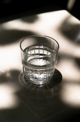 A clear glass of water sits on a reflective surface, illuminated by soft, dappled light and shadows.