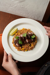 Hands hold a white plate with sliced, seared duck breast drizzled with a dark sauce, resting on fried rice with corn and cilantro, garnished with a lime wedge