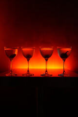 Four coupe cocktail glasses lined up on a dark, textured bar surface. Orange and red backlight, creating an atmospheric and sophisticated nightlife scene