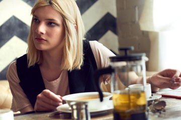 Transgender person in cafe. Close-up portrait of stylish blond transwoman sitting in cafe and looking away pensively