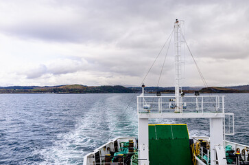 View from the back of a ferry crossing calm coastal waters over to the Isle of Mull in Scotland. Leaving a trail of waves under cloudy skies toward distant land.