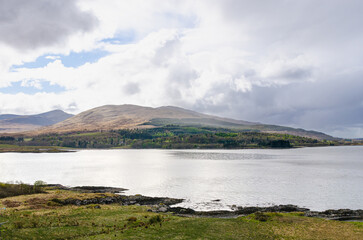 Isle of Mull - Beautiful view of a calm lake with rolling hills and mountains in the background under soft sunlight and dramatic clouds.
