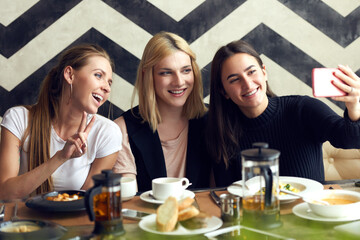 Friends meeting in cafe. Young pretty girls and their transgender friend sitting at table in cafe, taking selfie on smartphone and smiling cheerfully