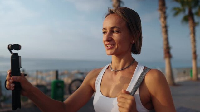 Fitness blogger woman films herself while walking outdoors on a sunny morning. She holds a camera in one hand and carries a yoga mat over her shoulder. Concept of active lifestyle and content creation