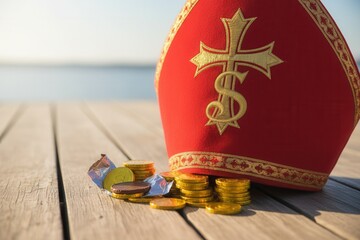 A red Sinterklaas mitre hat and chocolate gold coins on a wooden pier. Traditional Dutch Saint Nicholas holiday celebration concept with copy space