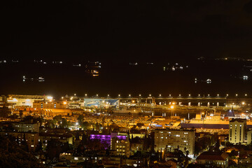 Distant view of docked ships and lit industrial harbor at night, surrounded by apartment buildings and city lights.