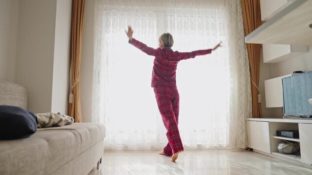 Young woman in red plaid pajamas opens the window, letting in fresh air. Her posture reflects relief and renewed energy. Concept of recovery, wellness, and fresh start. Back view, natural light, mid