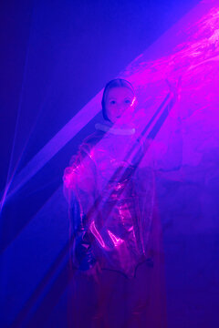 Vertical medium long studio portrait of senior Caucasian woman wearing unusual metallic outfit posing on camera in violet, blue and pink neon light with smoke and liquid texture effect, generation Z f