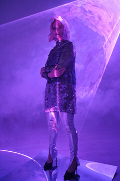 Vertical full body shot of senior woman wearing stylish metallic sweater and sequin dress above it with corrugated tube on head standing in neon light