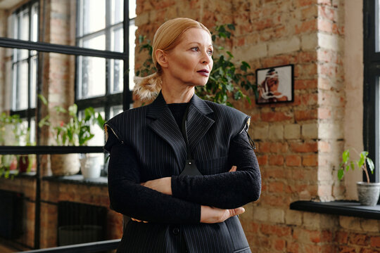 Medium portrait of confident senior Caucasian woman wearing modern black goth outfit standing in loft office with arms crossed looking away