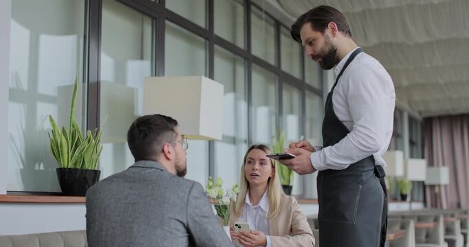 Waiter takes an order from a young couple in restaurant. Cafe worker taking note, writing order. Customer service.