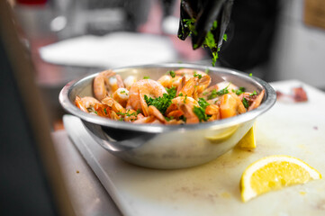 Close-up of cooked shrimp in a metal bowl, garnished with fresh herbs by a gloved hand, with lemon wedge on cutting board in professional kitchen setting.
