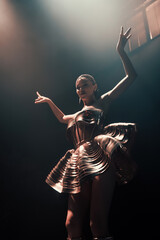 Go-go dancer in sculptural metallic dress posing on nightclub stage with arms raised. Warm spotlight and haze create dramatic, glamorous mood. Vertical photo.
