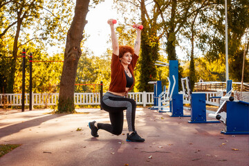 Adult Caucasian woman wearing sport clothes training in city park. Athlete does squats with dumbbells in her hands