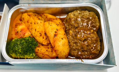 Airline meal featuring two beef rissoles smothered in gravy, served alongside seasoned potato wedges, a piece of broccoli, and a slice of tomato in a foil tray.