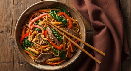 Overhead view of a delicious stirfry noodle dish with beef, vegetables, and chopsticks in a rustic bowl on a wooden table